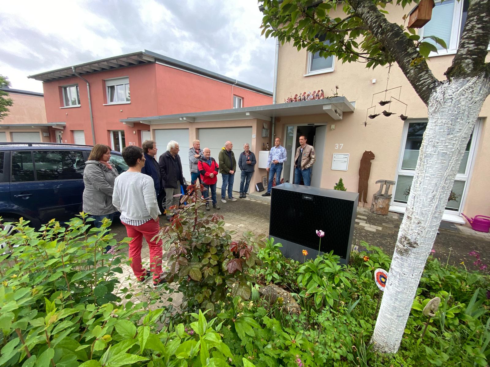 Eine Menschengruppe steht vor einem Haus und diskutiert. Im Vordergrund ist die im Vorgarten platzierte Außeneinheit einer Wärmepumpe zu sehen.