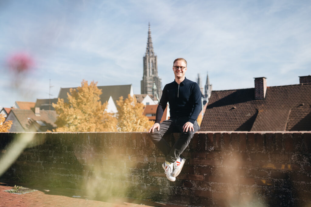 Marcel Emmerich sitzt auf der Ulmer Stadtmauer, das Münster ist im Hintergrund zu sehen.