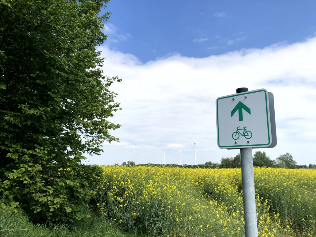 Rapsfeld mit einem Radwegschild im Vordergrund und mehreren Windkraftanlagen im Hintergrund